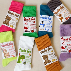 Könnte beinhalten: Verschiedene bunte Socken mit Geschenkanhängern. Jede Socke hat ein anderes Design, darunter ein Bär, der Weihnachtsmann, ein Pinguin, ein Rentier, ein Hase, ein Dinosaurier und ein Teddybär. Die Anhänger tragen die Aufschrift "Danke" und enthalten Geburtstagsdetails.