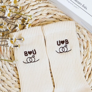 Può includere: Un paio di calzini color crema con ricami di testo e disegni di anelli. Un calzino recita "B♥U" e l'altro "U♥B". I calzini sono piegati su una superficie tessuta, con fiori bianchi sullo sfondo.