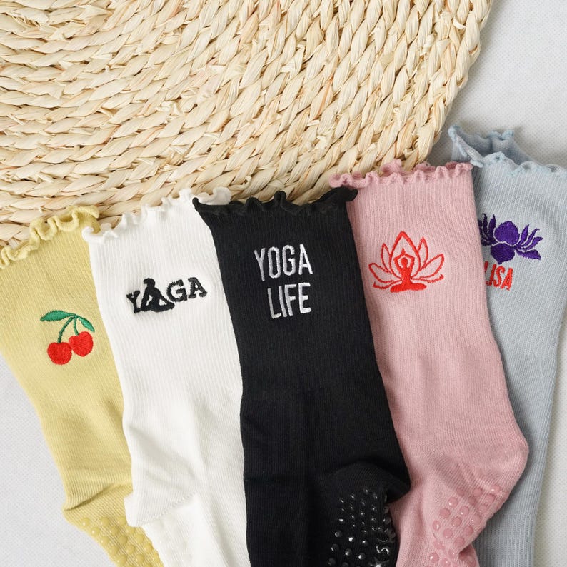 May include: Five pairs of colourful socks with different embroidered designs. The socks have a frilly edge and grippy dots on the bottom. The designs include a cherry, the word "YOGA", a lotus flower, the word "YOGA LIFE", and a flower.