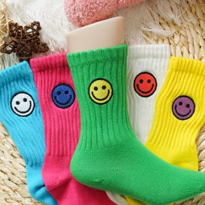 Op de afbeelding: Een close-up van vijf kleurrijke sokken, elk met een andere kleur en een smiley-ontwerp. De sokken zijn blauw, roze, groen, wit en geel. De smileys zijn zwart en de sokken zijn geribbeld.