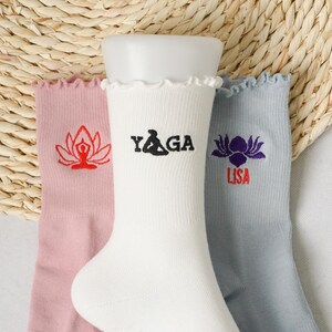 May include: Three pairs of white cotton socks with embroidered designs. The socks have a frilled top. One pair has a red lotus flower with a person meditating inside, another pair has the word "YAGA" in black lettering, and the last pair has a purple lotus flower with the word "LISA" in red lettering.
