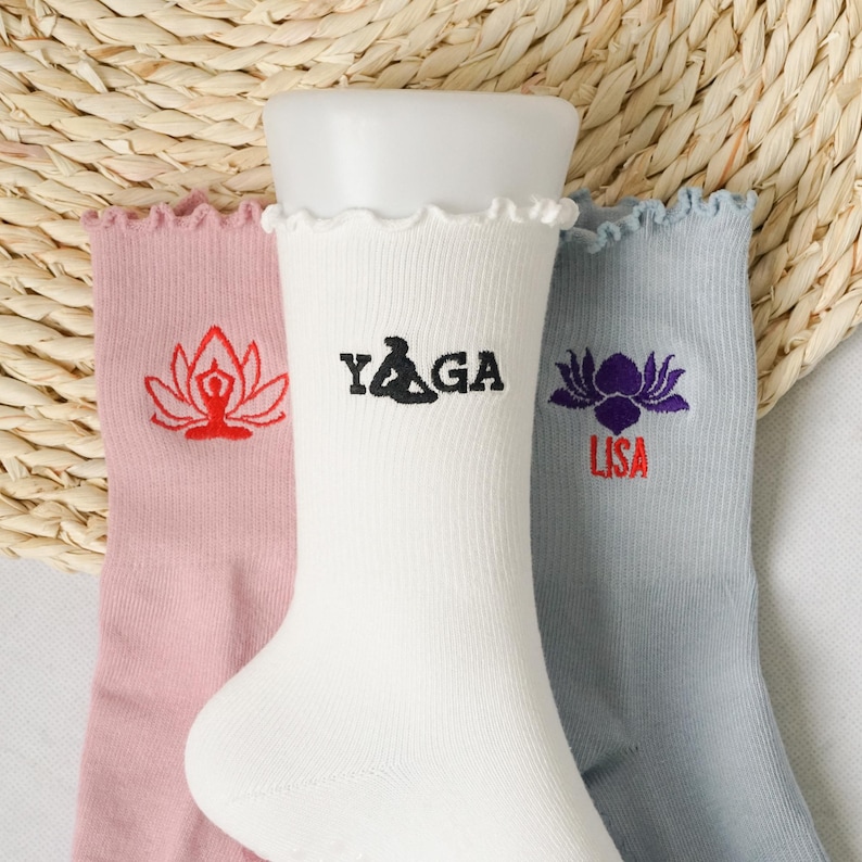 May include: Three pairs of white cotton socks with embroidered designs. The socks have a frilled top. One pair has a red lotus flower with a person meditating inside, another pair has the word "YAGA" in black lettering, and the last pair has a purple lotus flower with the word "LISA" in red lettering.
