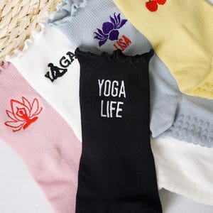May include: A collection of five pairs of socks with different designs. The socks are all white with different coloured accents and designs. One pair has a black background with the text "YOGA LIFE" in white. Another pair has a pink background with a red lotus flower design. Another pair has a white background with a black yoga pose design. Another pair has a light blue background with a purple lotus flower design. The last pair has a yellow background with a red cherry design.