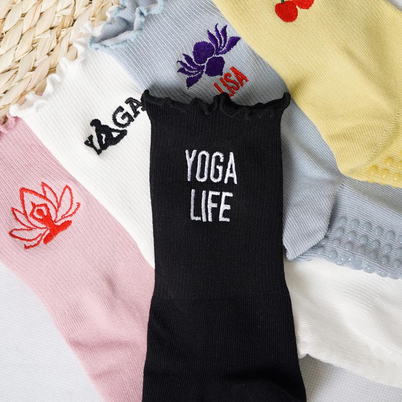 May include: A collection of five pairs of socks with different designs. The socks are all white with different coloured accents and designs. One pair has a black background with the text "YOGA LIFE" in white. Another pair has a pink background with a red lotus flower design. Another pair has a white background with a black yoga pose design. Another pair has a light blue background with a purple lotus flower design. The last pair has a yellow background with a red cherry design.