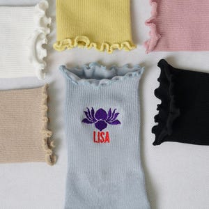 May include: Five pairs of ribbed socks with frilly edges. The socks are in various colours, including white, yellow, pink, beige, and black. One pair of socks has a purple lotus flower and the name "LISA" embroidered on them.