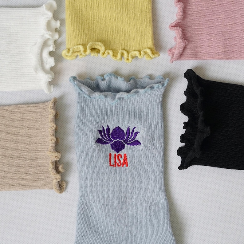 May include: Five pairs of ribbed socks with frilly edges. The socks are in various colours, including white, yellow, pink, beige, and black. One pair of socks has a purple lotus flower and the name "LISA" embroidered on them.