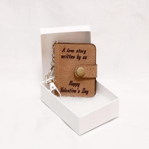 May include: A brown leather keychain with a small book charm. The book charm has the text "A love story written by us" and "Happy Valentine's Day" engraved on it. The keychain is in a white box.
