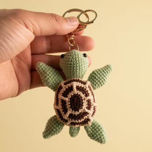 May include: A crocheted turtle keyring in shades of green and brown, with a gold-coloured keyring. The turtle has a detailed shell pattern and small black eyes. The keyring is held up against a light yellow background.