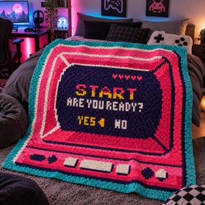 May include: A crochet blanket featuring a pixelated retro video game console screen design in vibrant pink, dark blue, and white. The screen displays the text START ARE YOU READY? with YES and NO options, mimicking an old-school gaming interface.