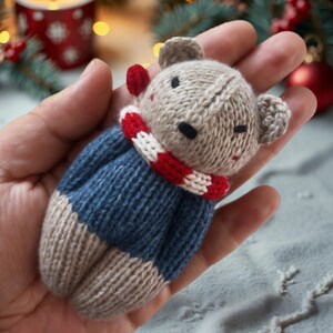 May include: A knitted gray and blue teddy bear wearing a red and white striped scarf. The bear is being held in a hand.