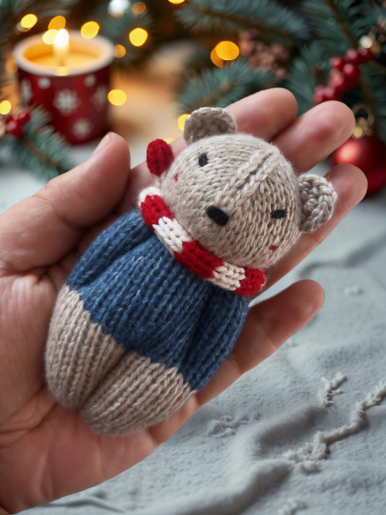 May include: A knitted gray and blue teddy bear wearing a red and white striped scarf. The bear is being held in a hand.