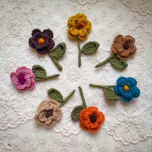 May include: Seven crocheted flower embellishments with green stems and leaves. The flowers are in various colors, including brown, pink, orange, yellow, and blue.