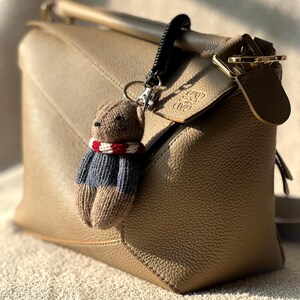 May include: A tan leather handbag with a brown knitted bear keychain. The bear is wearing a red and white striped scarf and a blue and white striped sweater.