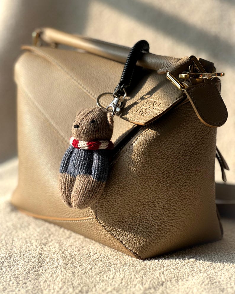 May include: A tan leather handbag with a brown knitted bear keychain. The bear is wearing a red and white striped scarf and a blue and white striped sweater.
