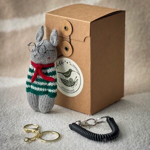May include: A gray knitted rabbit wearing glasses and a green and white striped sweater with a red scarf. The rabbit is holding a key chain with a black spring and a silver clip. The rabbit is sitting on a brown surface next to a brown box with a label that says "weaversnest".