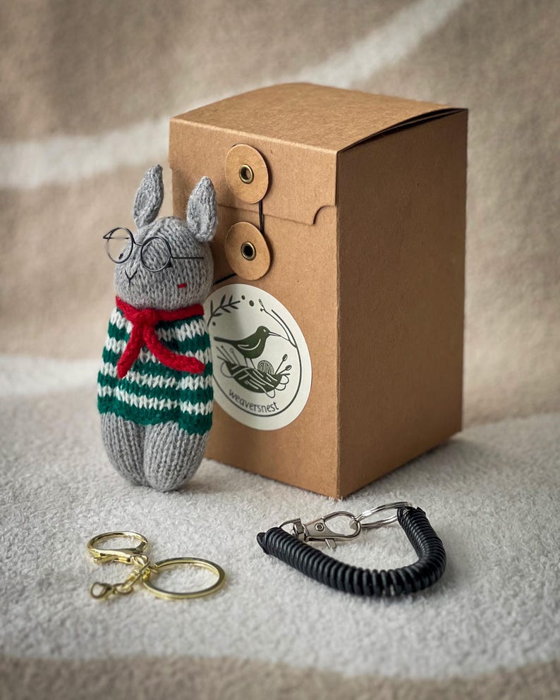 May include: A gray knitted rabbit wearing glasses and a green and white striped sweater with a red scarf. The rabbit is holding a key chain with a black spring and a silver clip. The rabbit is sitting on a brown surface next to a brown box with a label that says "weaversnest".