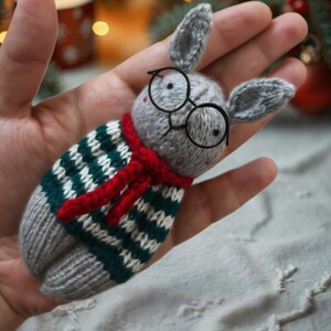 May include: A gray knitted bunny wearing a red and green striped sweater and black glasses. The bunny is being held in a hand.