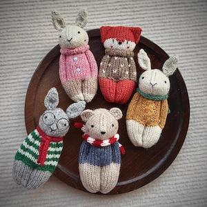 Peut inclure: Cinq jouets en peluche d'animaux sur une assiette en bois. Les jouets sont un lapin portant un pull rose, un renard portant un pull marron, un lapin portant un pull jaune, un ours portant un pull bleu et un lapin portant un pull rayé vert et rouge.