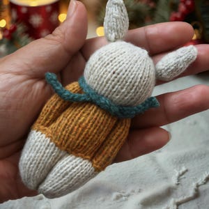 May include: A hand holding a knitted white bunny wearing a mustard yellow sweater with a teal green scarf.