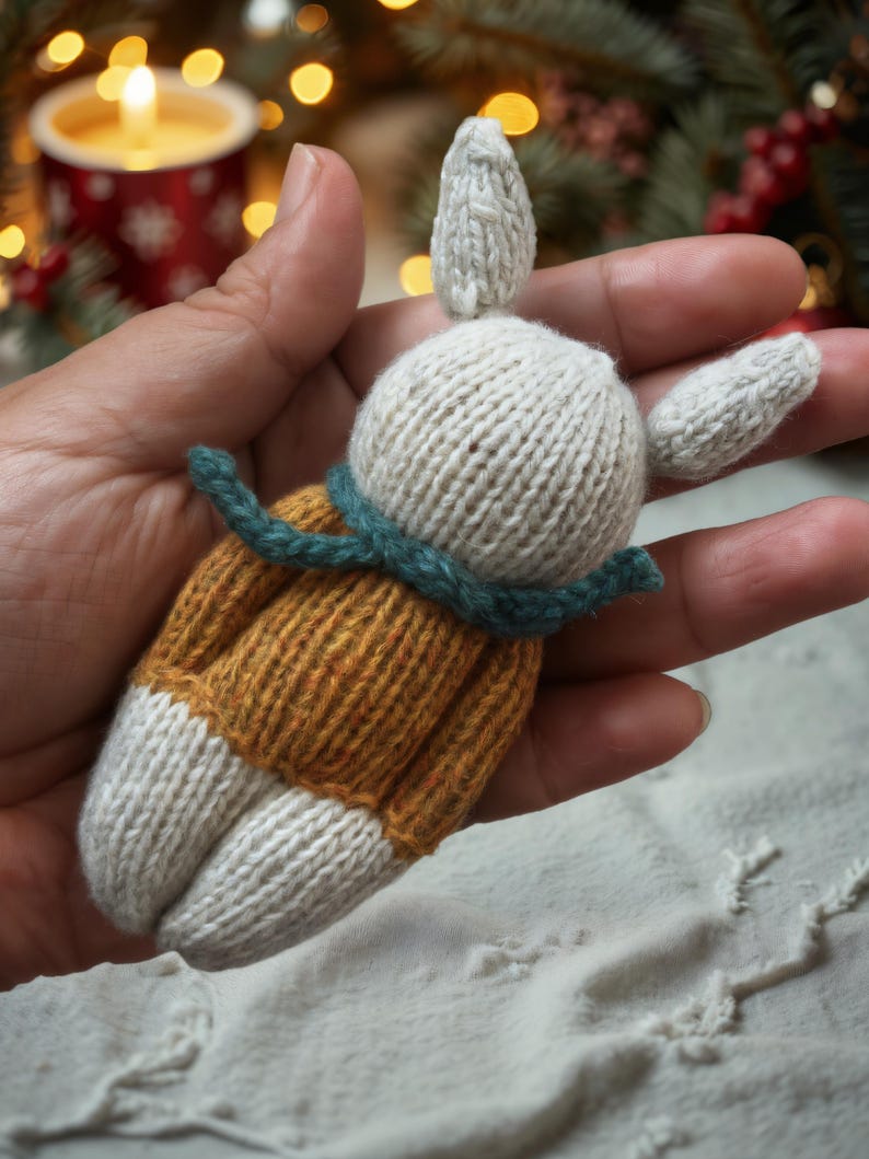 May include: A hand holding a knitted white bunny wearing a mustard yellow sweater with a teal green scarf.