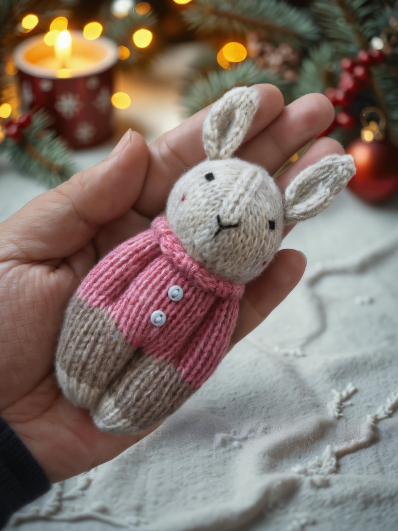 May include: A knitted bunny toy wearing a pink sweater with white buttons. The bunny is held in a hand against a white background.