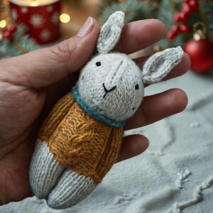 May include: A hand holding a knitted white bunny wearing a yellow sweater with a cable knit pattern. The bunny has black eyes and a blue scarf.