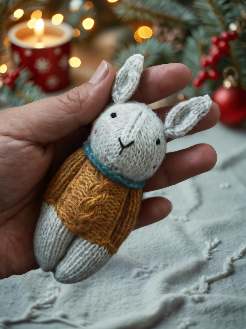 May include: A hand holding a knitted white bunny wearing a yellow sweater with a cable knit pattern. The bunny has black eyes and a blue scarf.