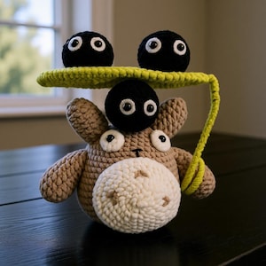 May include: A handcrafted crochet toy depicting a light brown cow-like creature with a cream-coloured muzzle. Three black, round soot sprites with white eyes are positioned above, with a lime green leaf-like structure above the cow's head.