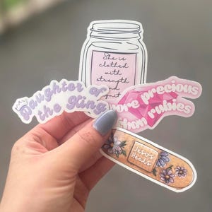 May include: A collection of stickers with inspirational quotes and designs. One sticker features a mason jar with the text "She is clothed with strength and grit." Other stickers include "Daughter of the King" and "more precious than rubies."