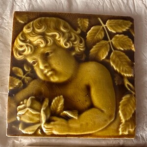 May include: Square ceramic tile with a relief of a cherub holding a rose, surrounded by leaves. The tile is primarily yellow-brown with darker brown accents, and has a crackled glaze finish. The cherub's curly hair and facial features are detailed.