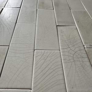 May include: Close-up of a floor tiled with rectangular, off-white tiles. Each tile has a unique pattern of fine, dark cracks, creating a textured, aged appearance. The tiles are arranged in a staggered, brick-like pattern.