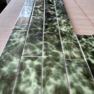 May include: A series of rectangular ceramic tiles arranged in a staggered pattern. Each tile features a glossy, crackled glaze with a mottled blend of dark forest green and pale sage green colours, creating a textured, marbled appearance.