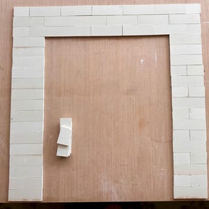 Puede incluir: Un marco rectangular construido con pequeños ladrillos rectangulares de color blanco roto dispuestos en un patrón de hiladas apiladas. Una pequeña pila de ladrillos idénticos sueltos se encuentra a la izquierda de la abertura central.