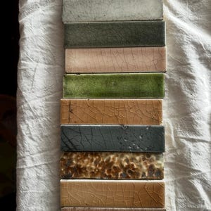 May include: A vertical stack of ten rectangular ceramic tiles, each featuring a distinct crackle glaze finish. The colors include shades of light green, off-white, dark olive, soft pink, moss green, tan, charcoal gray, mottled brown, and light brown.