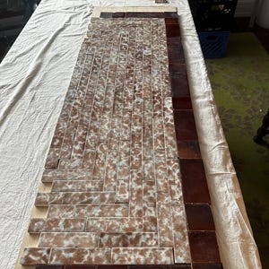 Puede incluir: Una disposición rectangular de azulejos sobre una tabla de madera. El centro presenta un patrón de espiga de azulejos marrones y blancos moteados, mientras que el borde consta de azulejos lisos de color marrón oscuro brillante.