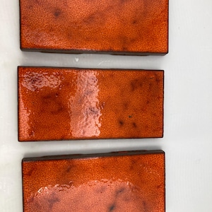 May include: Three rectangular orange tiles with a textured surface and dark brown edges. The tiles have a glossy finish, and are arranged vertically against a white background. The tiles are likely for home decor or construction.