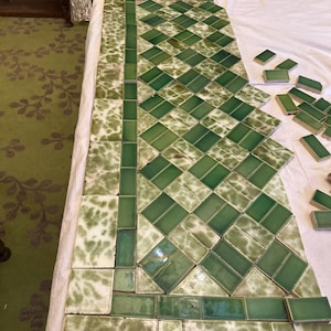 Puede incluir: Azulejos de cerámica verdes y blancos dispuestos en un patrón de diamantes, con un borde de azulejos rectangulares. Los azulejos tienen un acabado brillante y están colocados sobre una superficie blanca, con azulejos sueltos adicionales cerca.