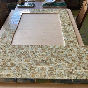 May include: A rectangular frame constructed from small, rectangular tiles in shades of tan, brown, and green. The tiles are arranged in a brick-like pattern, with a wooden surface visible within the frame. Additional tiles are scattered on the table.