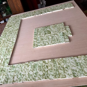 May include: A light-colored wooden surface with a square border and a smaller square in the center, both outlined with small, rectangular green and white tiles. The tiles have a speckled pattern.