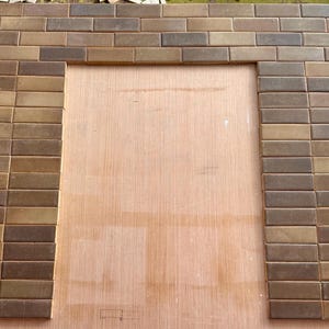 Puede incluir: Un marco rectangular construido con baldosas de cerámica rectangulares de color marrón y bronceado dispuestas en un patrón de ladrillo. Las baldosas están montadas sobre una tabla de madera plana y clara, creando un borde alrededor de un espacio vacío.