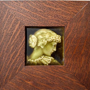 May include: A square ceramic tile featuring a relief profile of a person with curly hair in a bun, finished in a glossy olive green glaze with a fine crackle pattern, set within a dark brown wooden frame with a prominent wood grain texture.