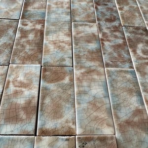 Puede incluir: Conjunto de baldosas cerámicas rectangulares con efecto craquelado. Las baldosas presentan una mezcla de tonos marrón, crema y azul claro, creando un aspecto texturizado y envejecido. Dispuestas en un patrón de rejilla.