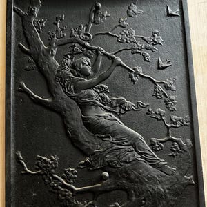 May include: A rectangular black cast iron fireback featuring a relief sculpture of a figure reclining in a tree. The figure reaches towards branches with leaves, while two small birds are depicted in flight in the upper right corner of the panel.