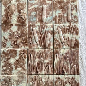May include: A collection of ceramic tiles with a brown and cream color scheme. Each tile features a relief design of aquatic plants, frogs, and lily pads. The tiles are arranged in a grid pattern, showcasing a nature-themed aesthetic.