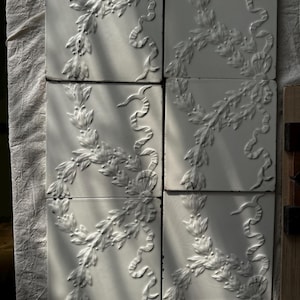 May include: A set of six square white ceramic tiles arranged in two columns. Each tile features a raised, embossed relief pattern of delicate floral garlands and flowing ribbons. The surface of the tiles has a glossy finish with visible fine cracks.