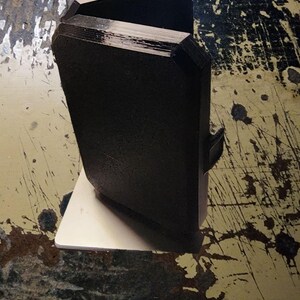 May include: A black, 3D printed phone stand with a rectangular base and a slightly angled top. The stand is designed to hold a phone upright.