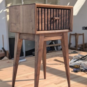 Può includere: Un comodino in stile moderno di metà secolo in legno scuro. Il comodino ha un singolo cassetto con doghe verticali e una piccola maniglia. Poggia su quattro gambe angolate. Dimensioni: circa 61 cm di altezza, 46 cm di larghezza e 41 cm di profondità.