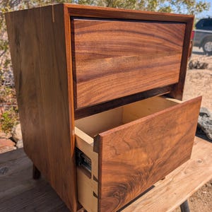 Pu&ograve; includere: Un comodino in legno marrone scuro con due cassetti caratterizzati da venature del legno visibili. Il cassetto superiore &egrave; chiuso, mentre quello inferiore &egrave; parzialmente aperto, rivelando fianchi interni chiari con giunzioni a coda di rondine.