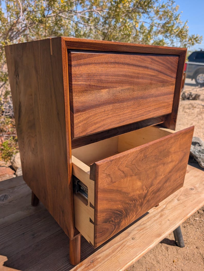 Pu&ograve; includere: Un comodino in legno marrone scuro con due cassetti caratterizzati da venature del legno visibili. Il cassetto superiore &egrave; chiuso, mentre quello inferiore &egrave; parzialmente aperto, rivelando fianchi interni chiari con giunzioni a coda di rondine.
