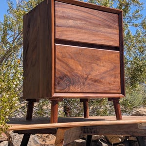 Pu&ograve; includere: Un comodino in legno marrone scuro con due cassetti sovrapposti caratterizzati da una venatura del legno naturale visibile. Il mobile &egrave; sostenuto da quattro gambe cilindriche corte e presenta una struttura rettangolare con bordi dritti.